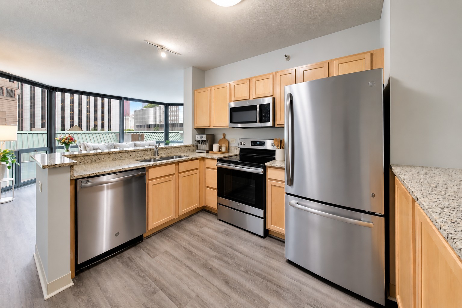 Condo kitchen with light wood cabinets, stainless steel appliances, and large windows overlooking city buildings.