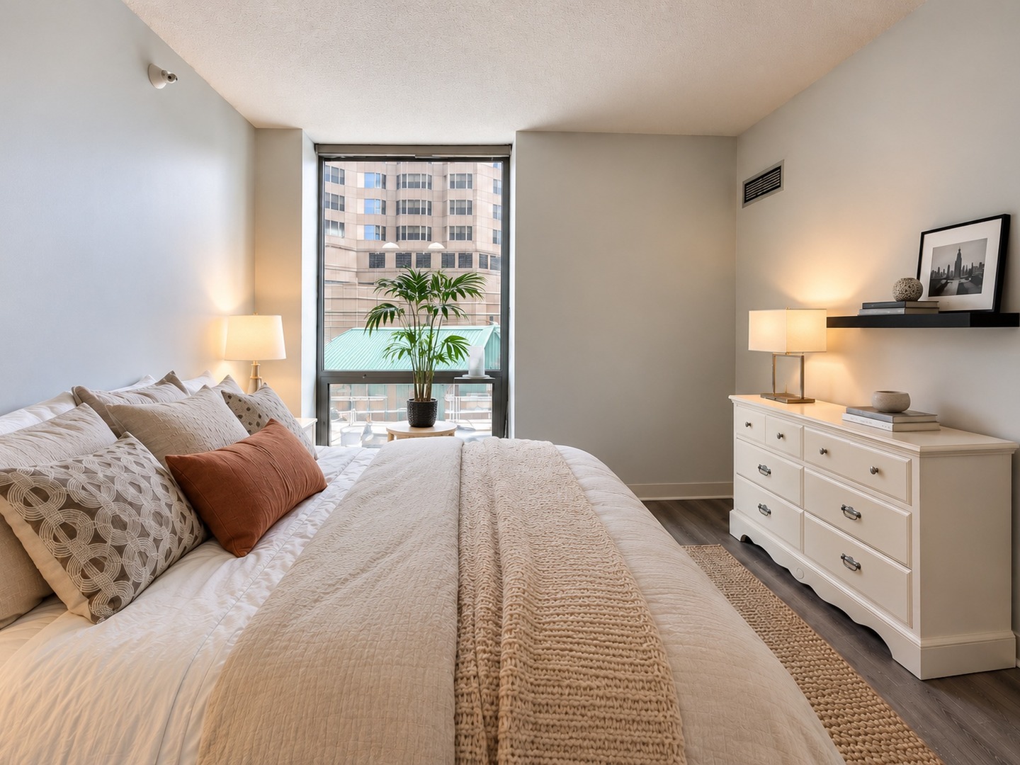 Staged bedroom with a white bed, rust accent pillow, dresser, and large window overlooking nearby buildings.