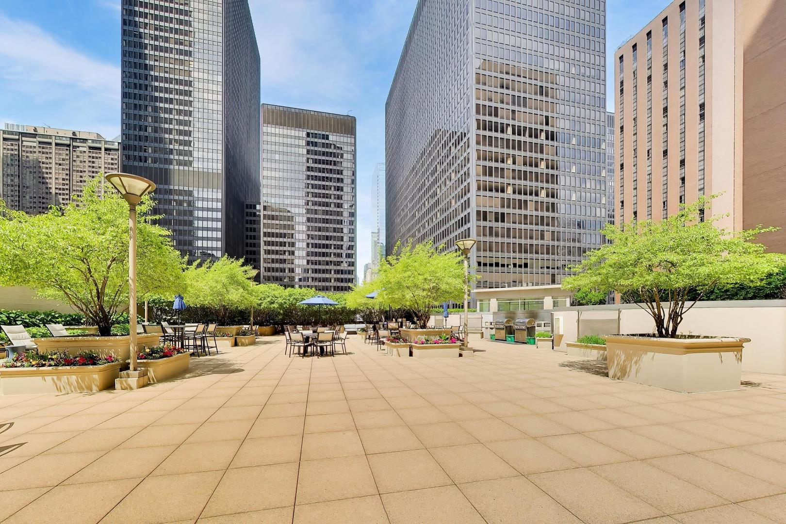 Rooftop terrace with outdoor seating, leafy planters, and surrounding high-rise buildings.