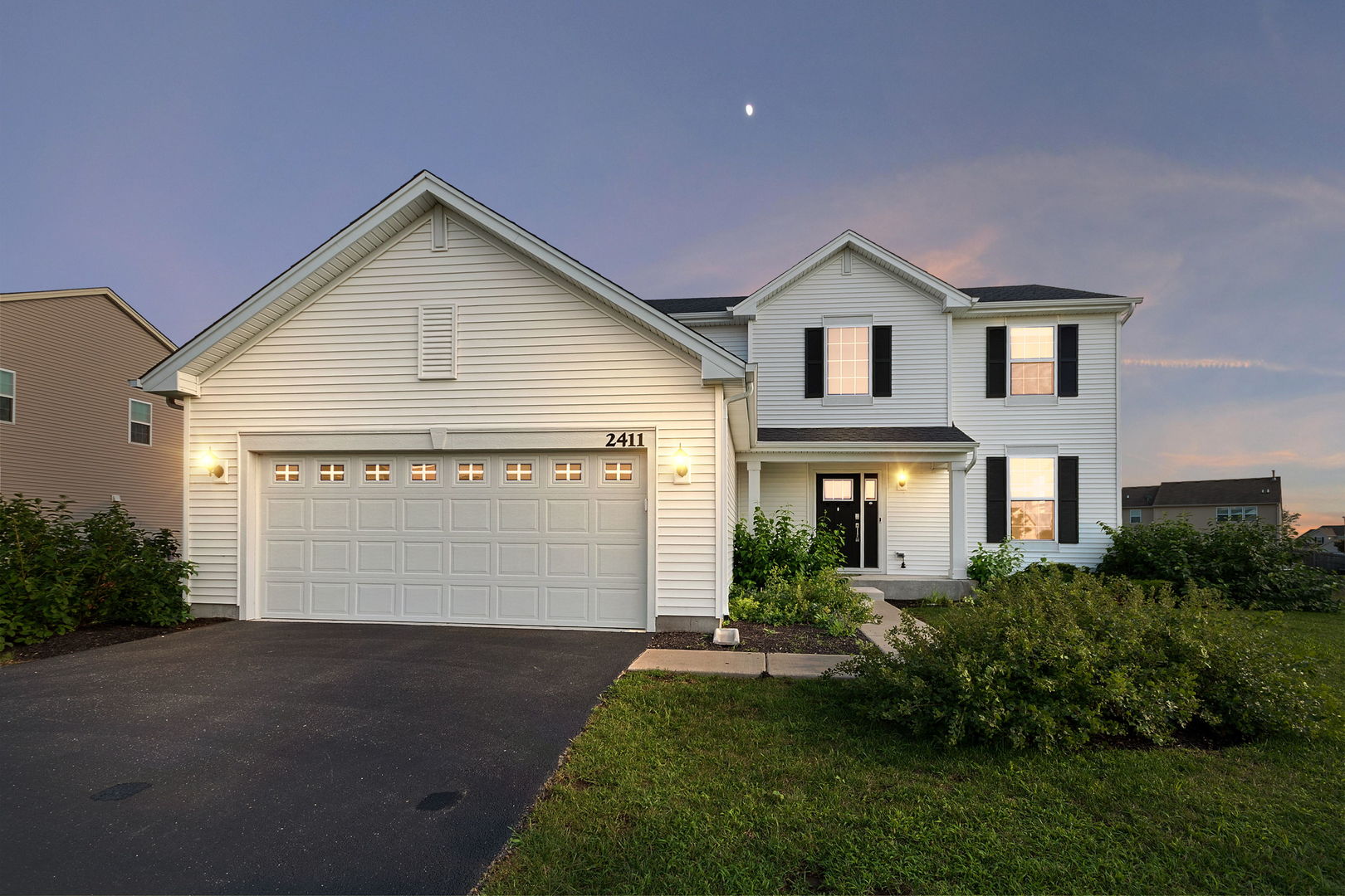 White two-story home with black shutters, an attached garage, and a landscaped front yard at dusk.