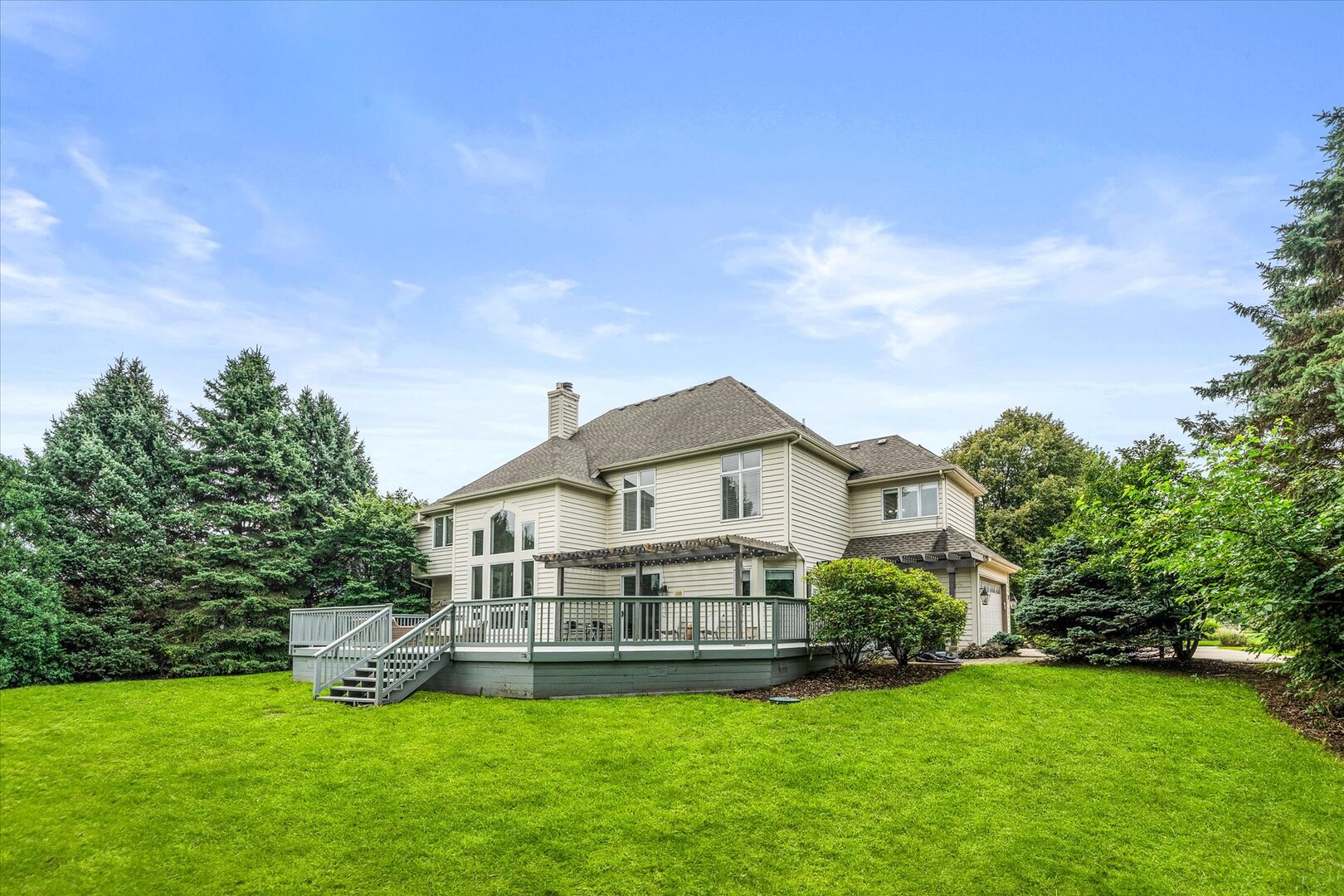 Two-story beige house with a raised deck and stairs overlooking a broad green lawn and mature trees.