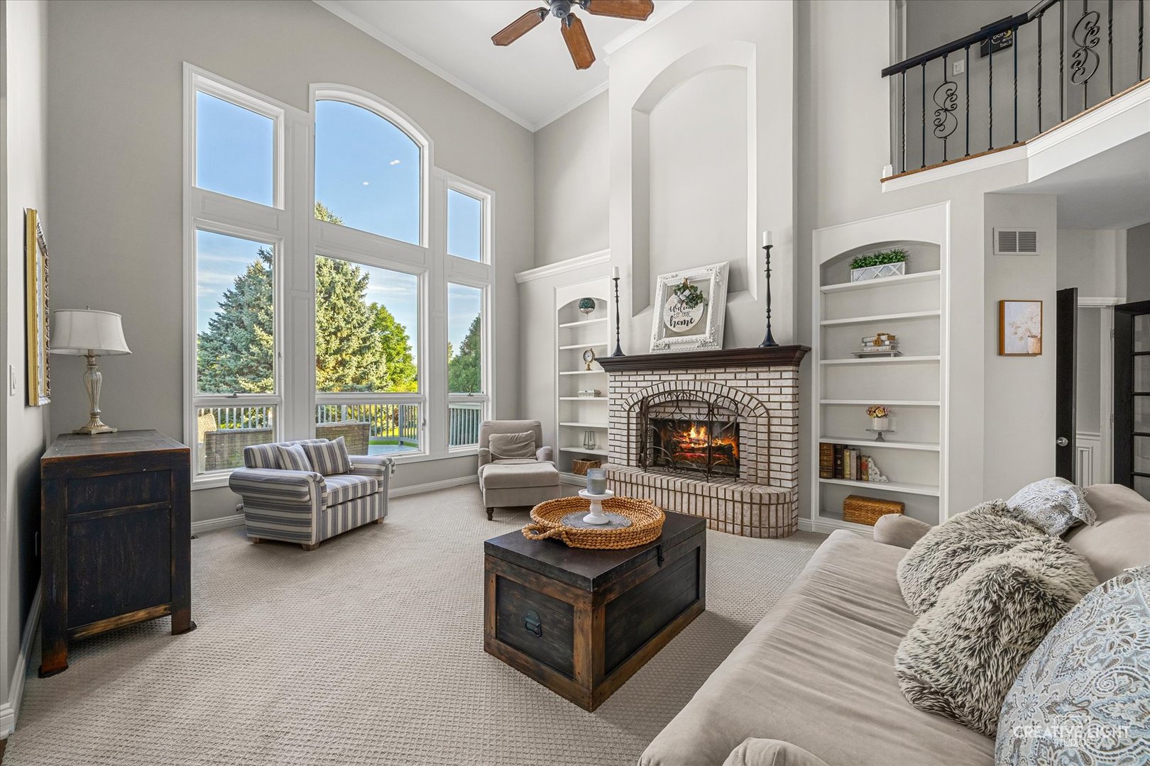 Living room with a tall brick fireplace, built-in shelves, and large windows overlooking greenery.