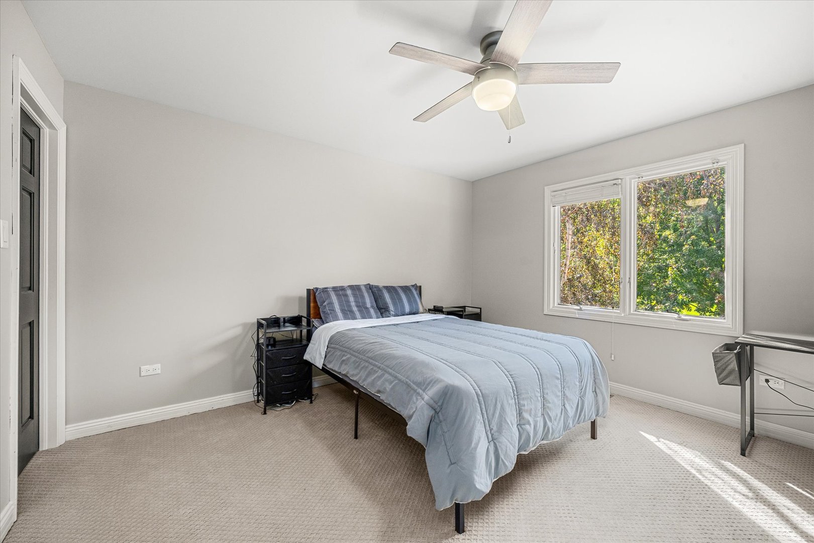 Bedroom with a pale blue bedspread, ceiling fan, and large window overlooking trees.