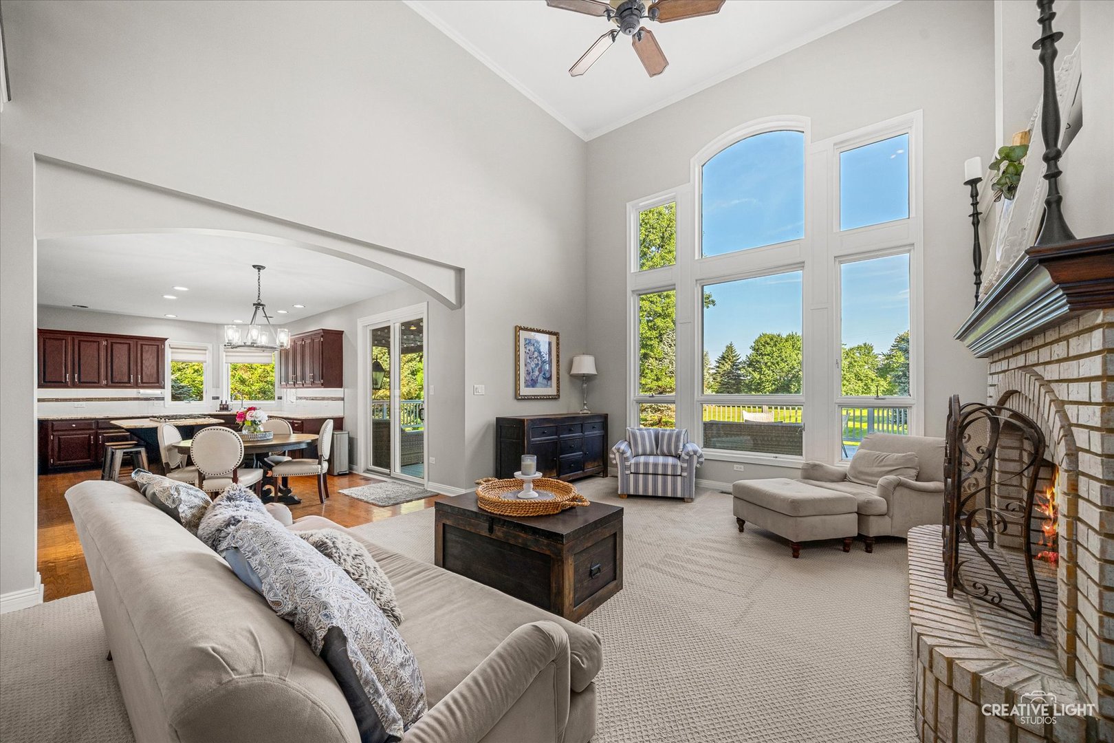 Living room with vaulted ceiling, tall windows, brick fireplace, and open dining area and kitchen.