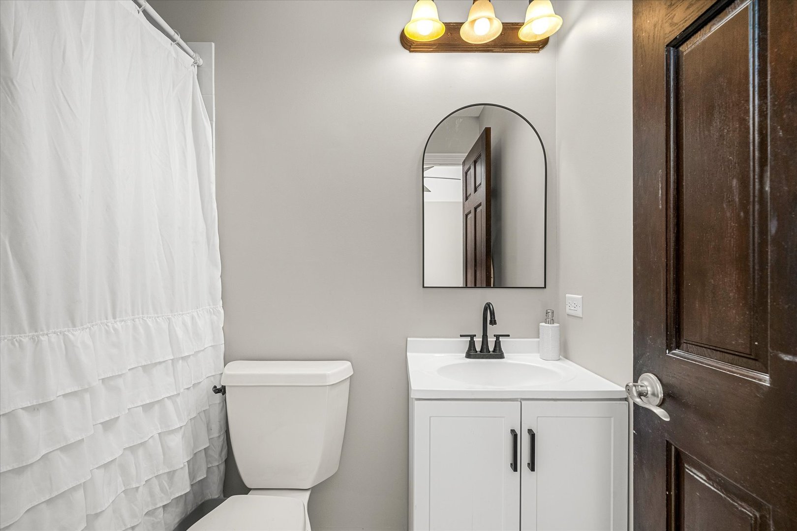 Bathroom with a white ruffled shower curtain, dark vanity fixtures, and an arched mirror.