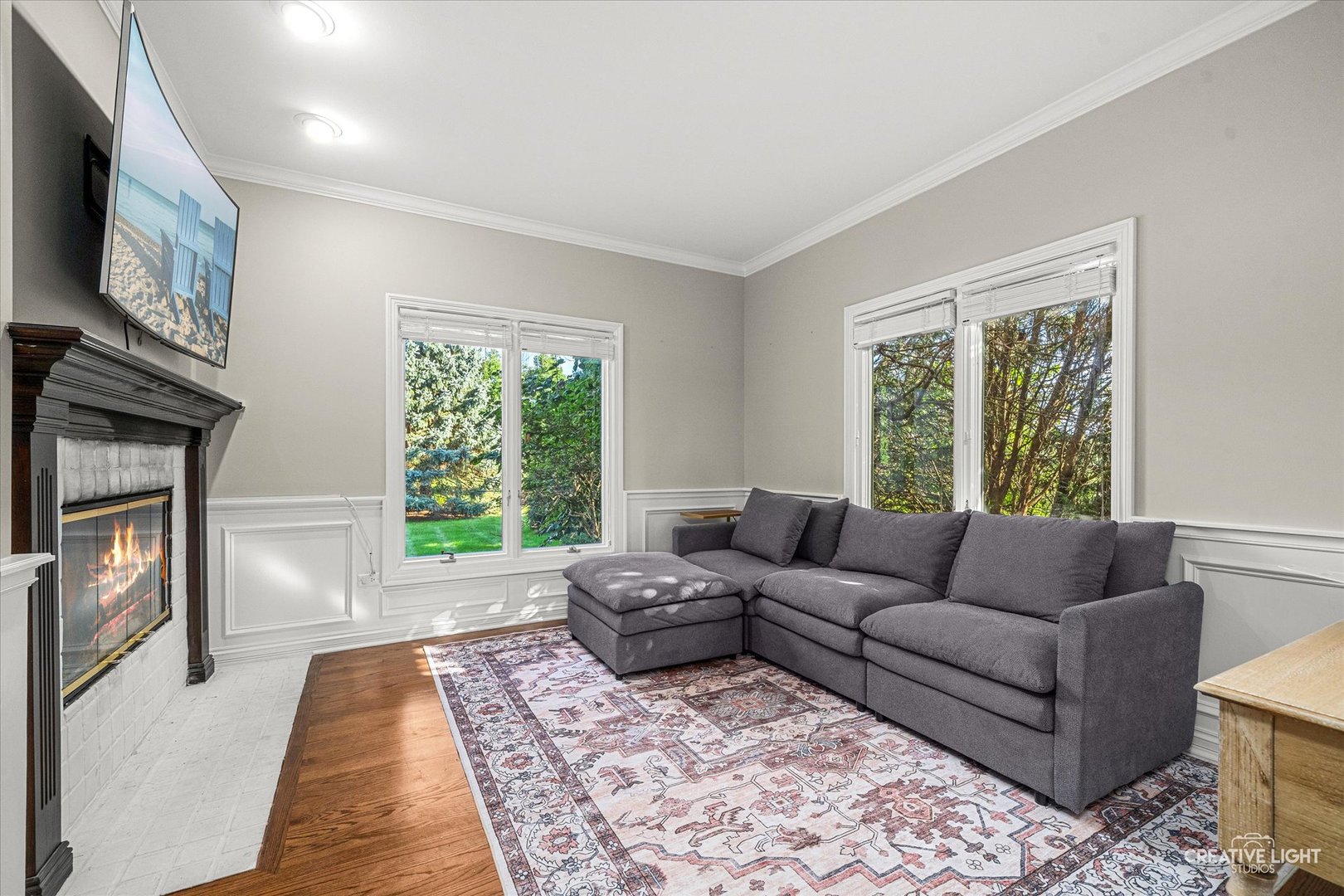 Living room with a gray sectional, patterned rug, large garden-facing windows, and a tiled fireplace.
