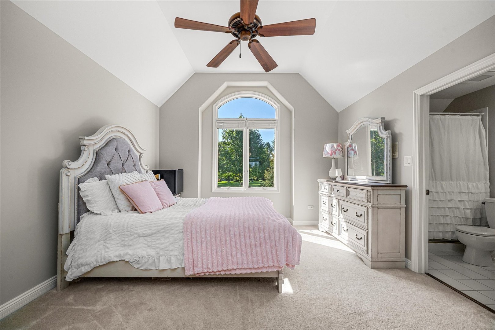 Bedroom with a vaulted ceiling, arched window, and white bed with a pink throw beside an ensuite bathroom.