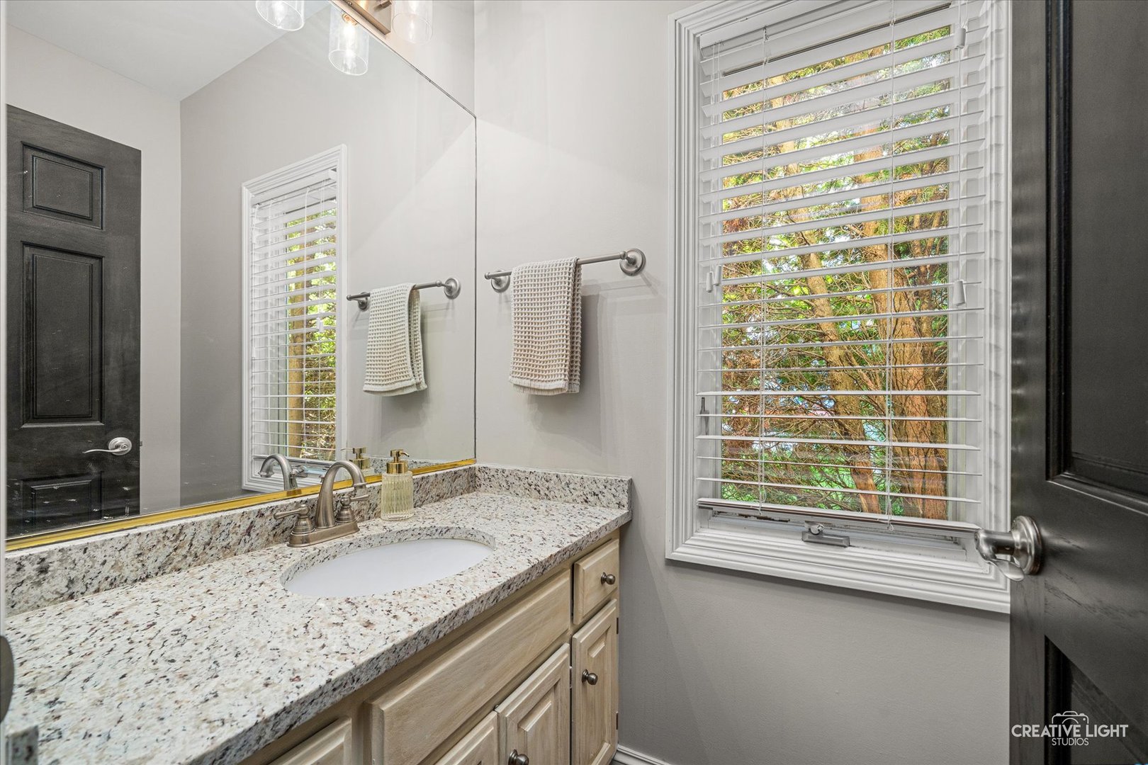Bathroom vanity with granite countertop, light wood cabinets, and a large window with white blinds.