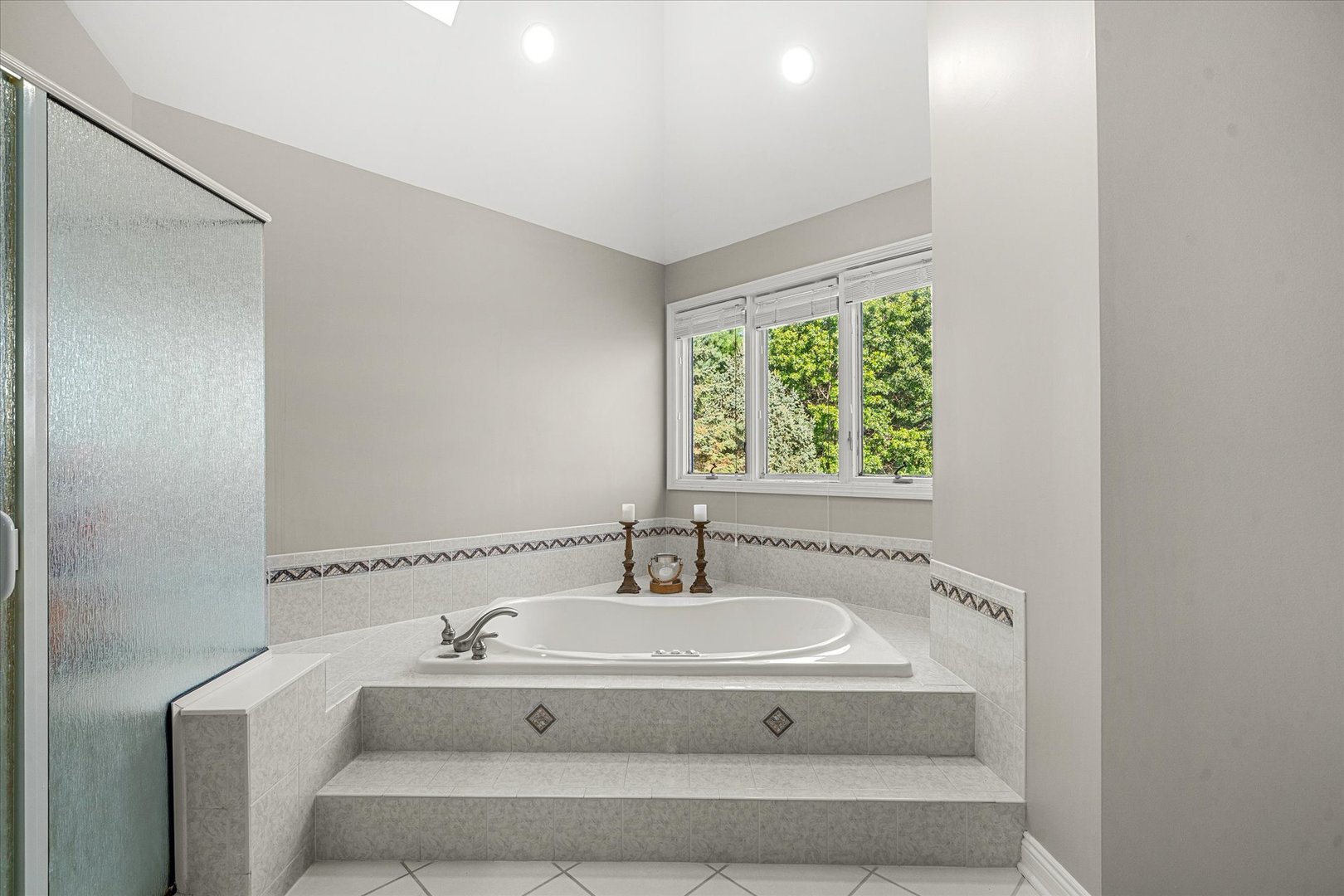 Bathroom with a corner soaking tub, tiled platform, frosted-glass shower, and window overlooking trees.