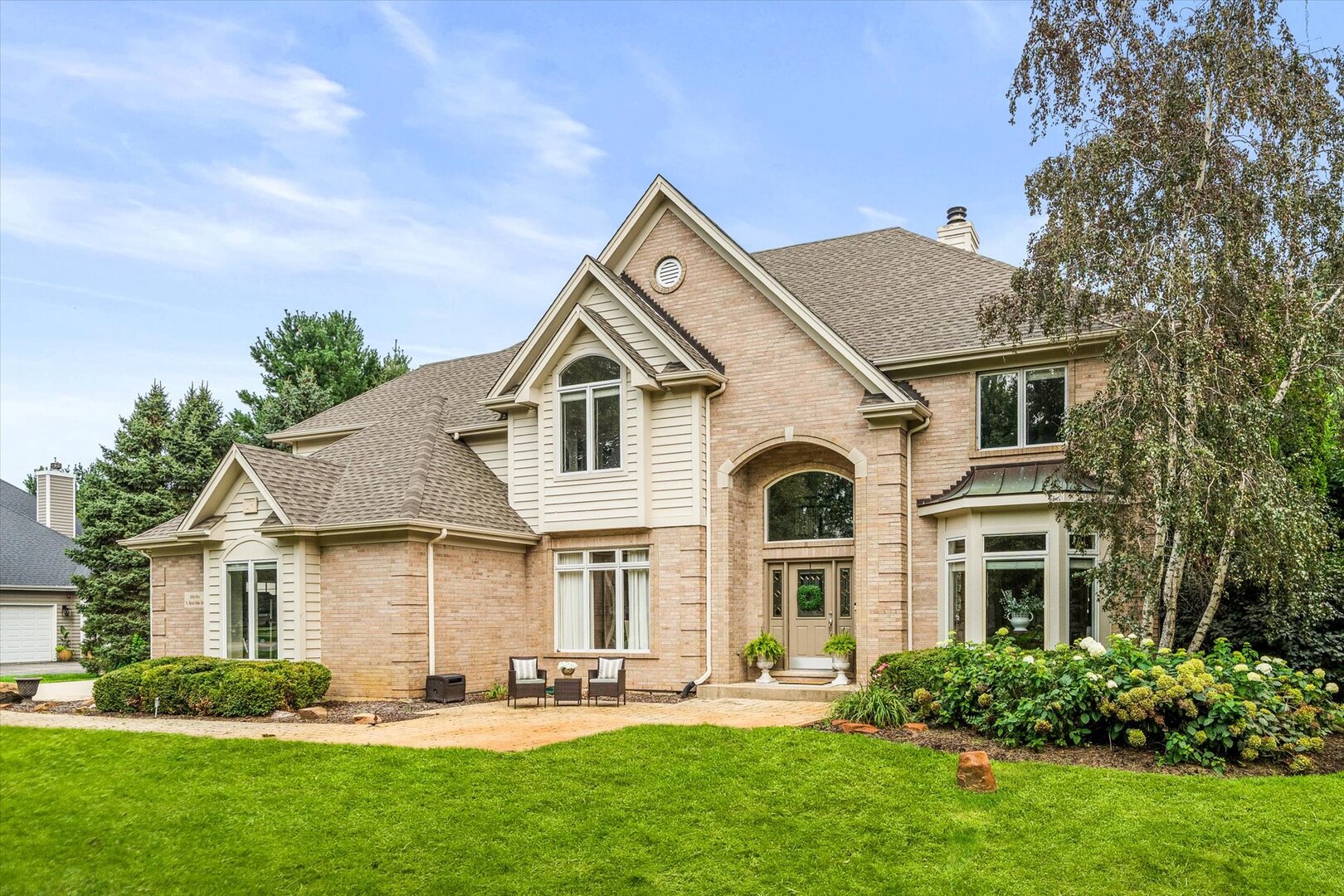 Two-story brick home with a gabled roof, bay window, landscaped lawn, and patio seating.