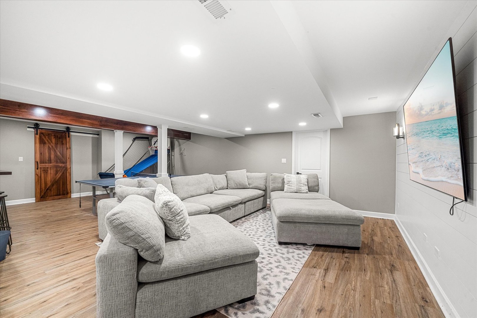 Basement family room with a gray sectional, wall-mounted TV, ping-pong table, and basketball arcade game.