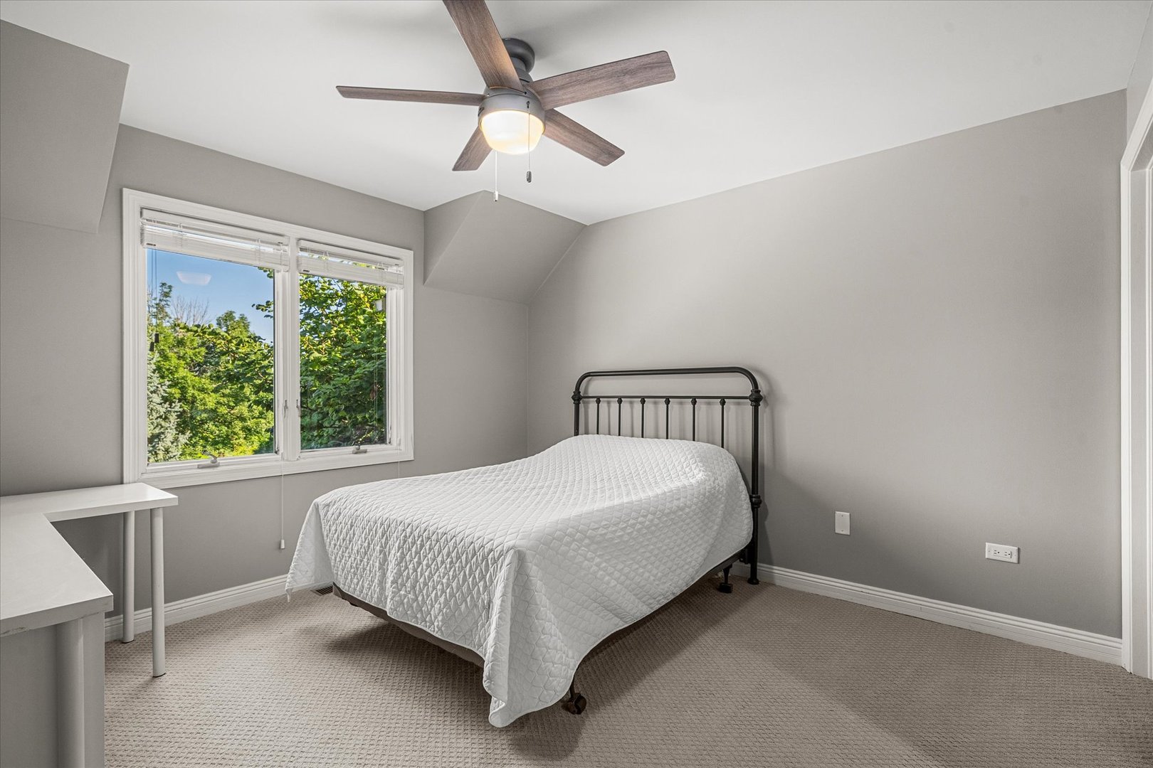 Bedroom with a white-quilted bed, ceiling fan, and window overlooking leafy greenery.