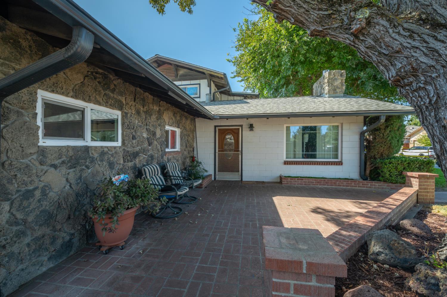 Home entrance with a covered brick patio, stone facade, rocking chairs, and a large tree.