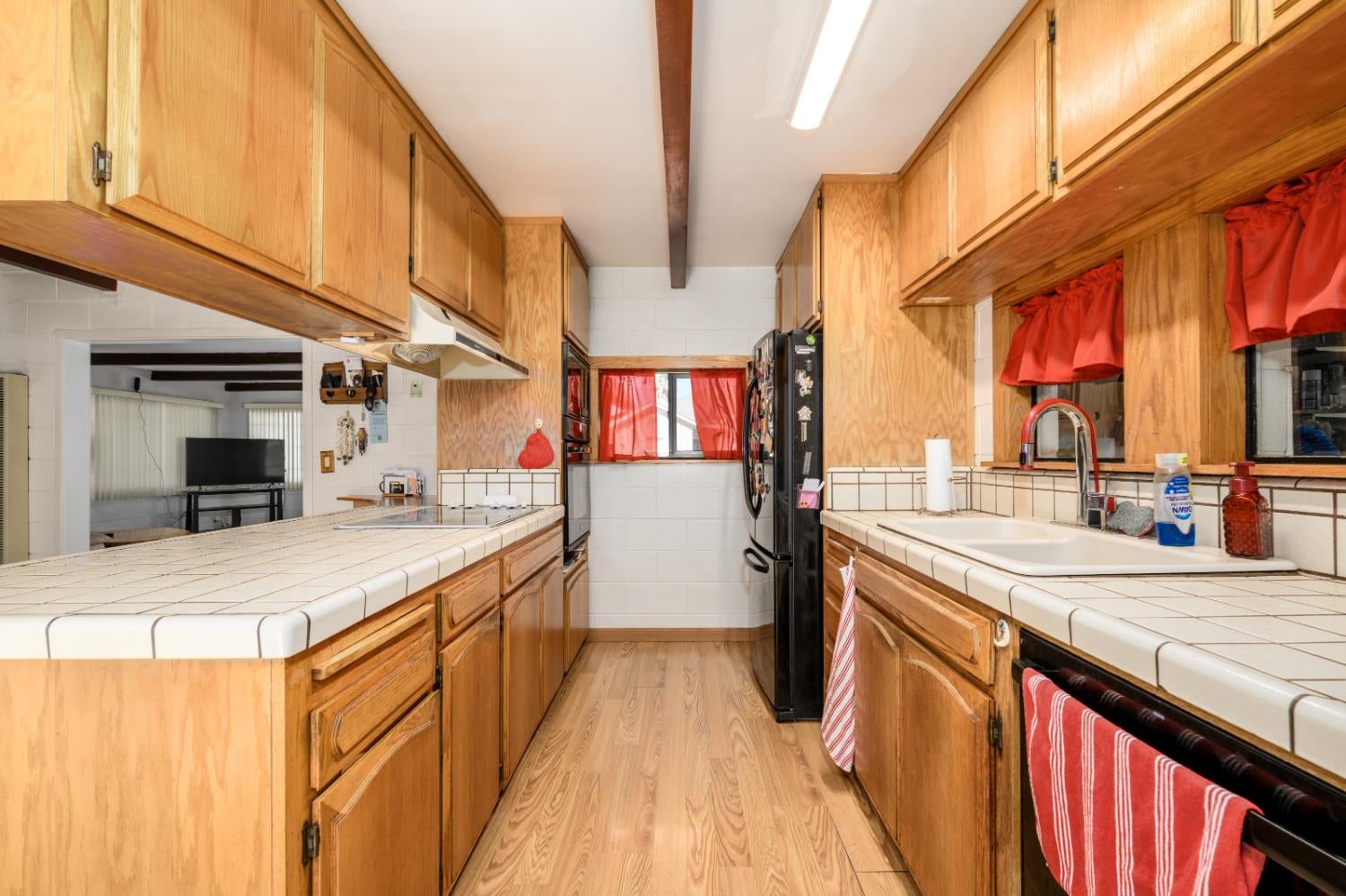 Galley kitchen with honey-colored cabinets, white tiled counters, and red window curtains.