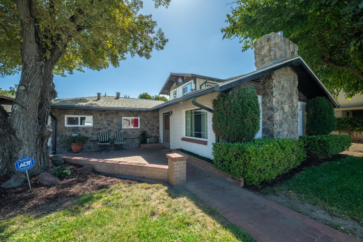 Stone-fronted house with a large chimney, brick patio, and mature trees.