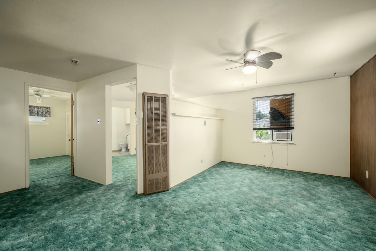 Empty room with green carpet, ceiling fan, wall heater, and window air conditioner.