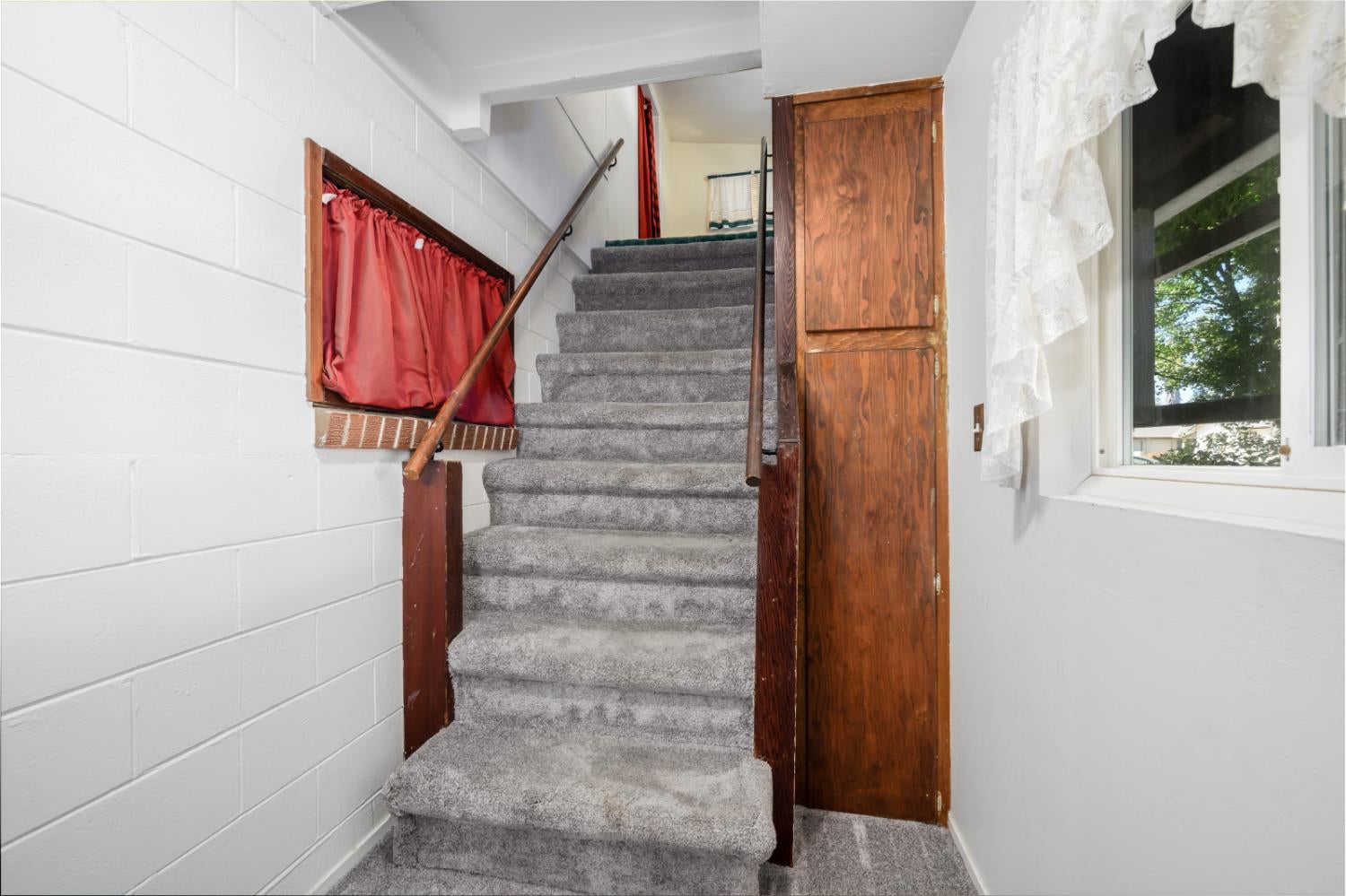 Carpeted staircase with dark wood handrails, red under-stair curtain, and a lace-curtained window.