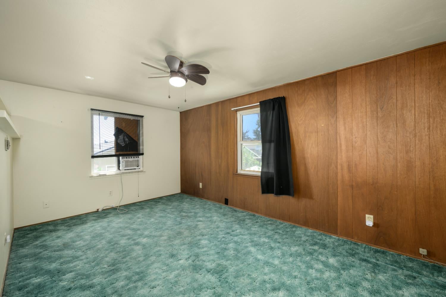 Empty room with teal carpet, wood-paneled walls, and a ceiling fan.