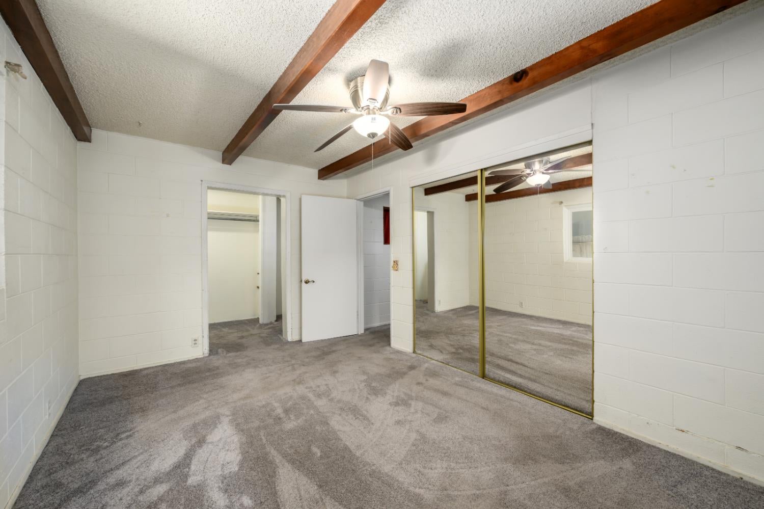 Empty bedroom with exposed ceiling beams, gray carpet, and mirrored sliding closet doors.
