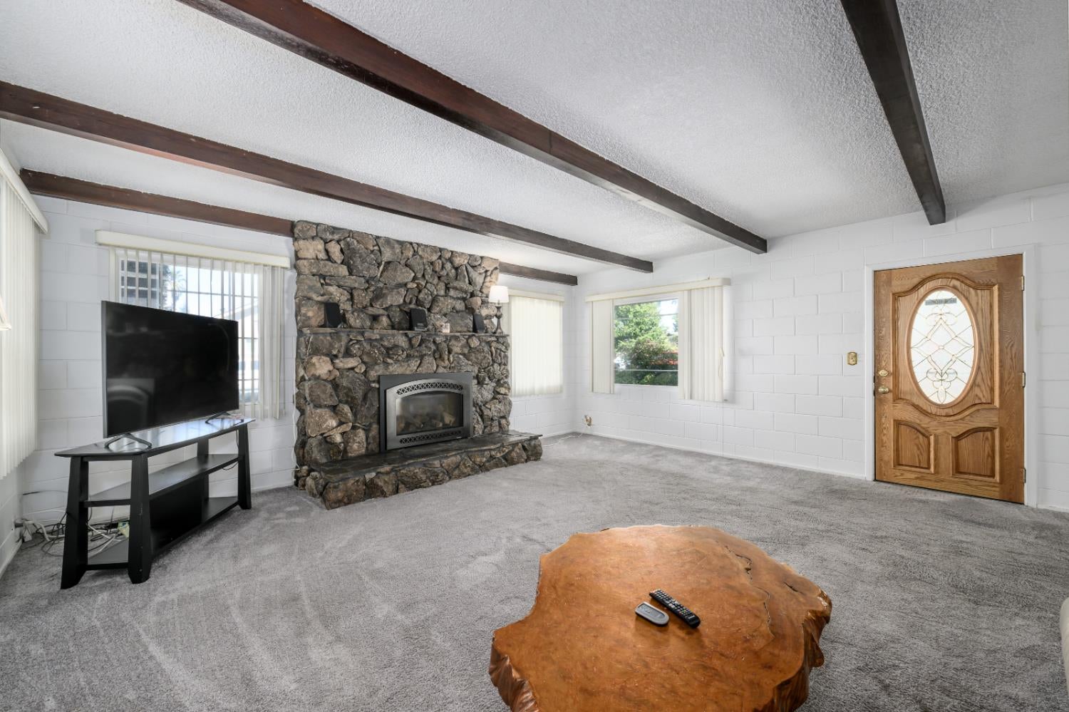Living room with a stone fireplace, exposed ceiling beams, gray carpet, and carved wooden front door.