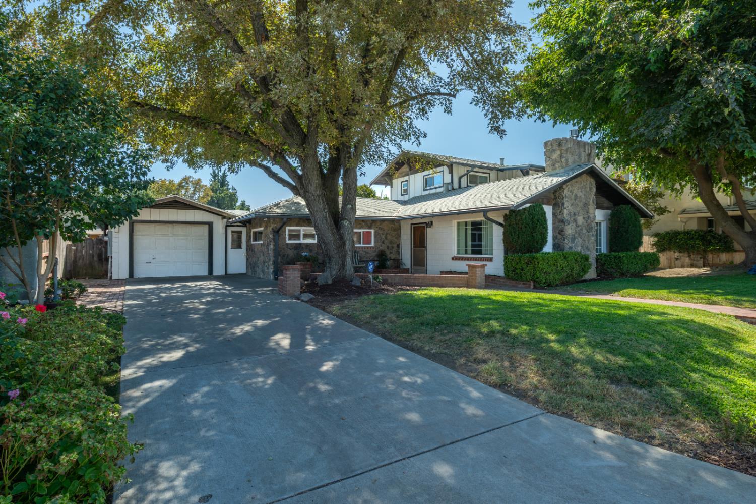Home with stone accents, attached garage, broad driveway, and a large shade tree in the front yard.