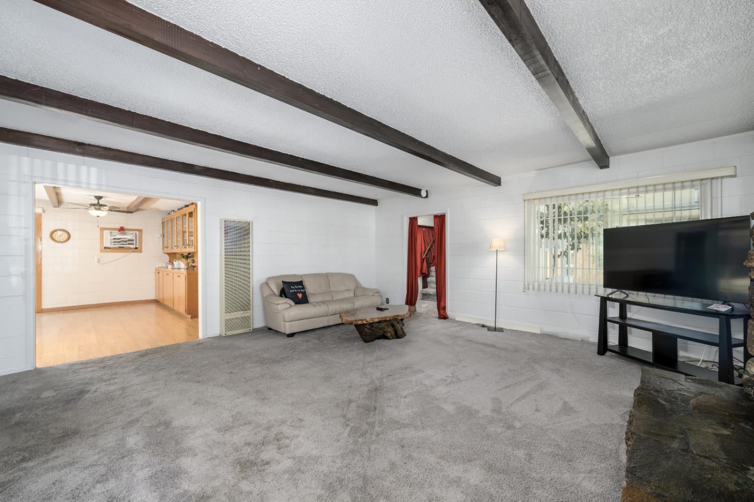 Living room with exposed ceiling beams, gray carpet, and a leather sofa beside an adjoining room.