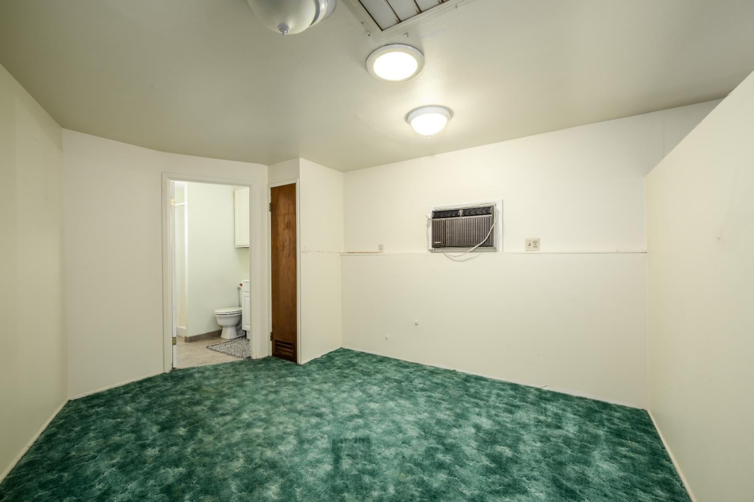 Empty carpeted room with a wall-mounted air conditioner and an open doorway to a bathroom.