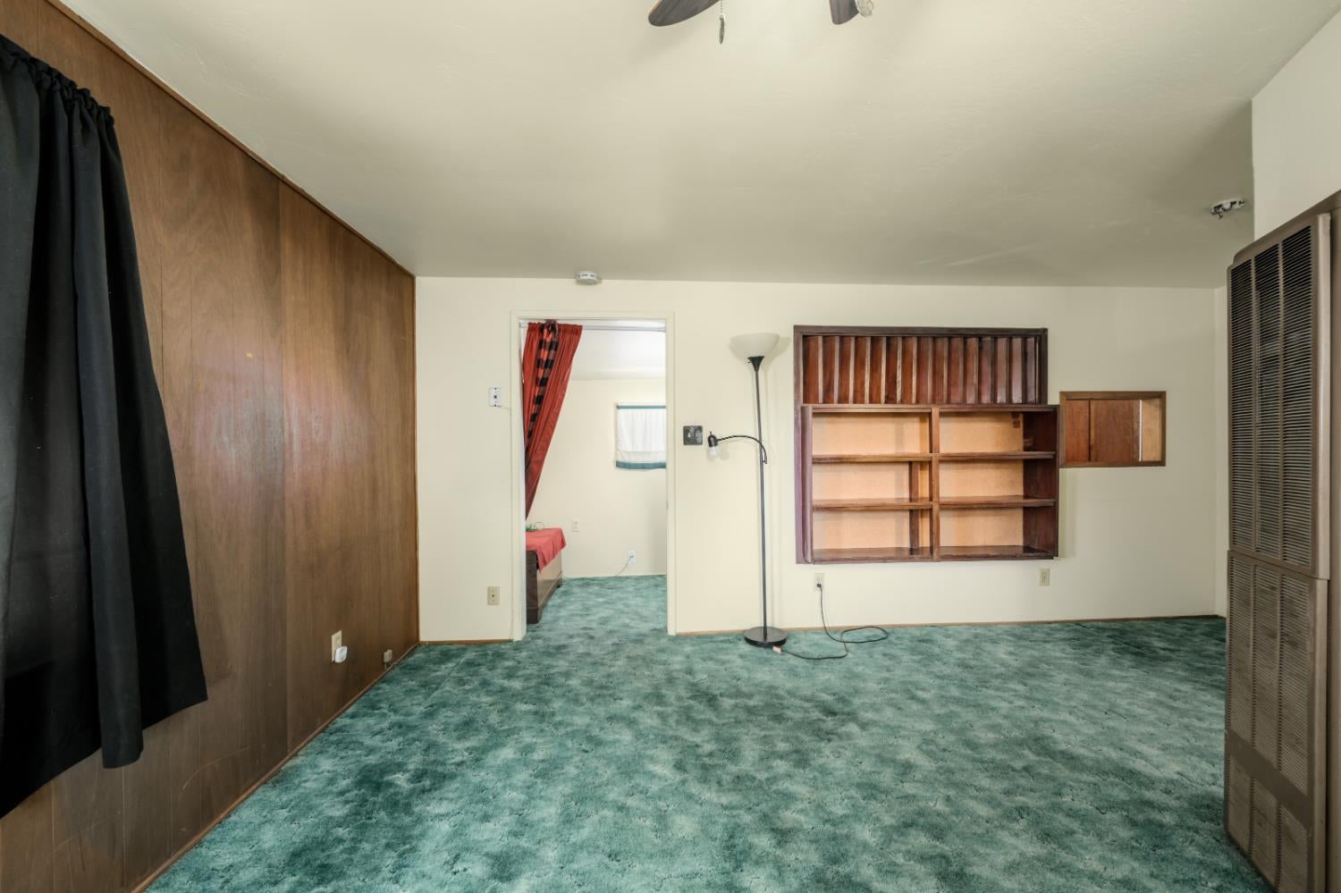 Mostly empty room with teal carpet, wood-paneled walls, and built-in wooden shelves.