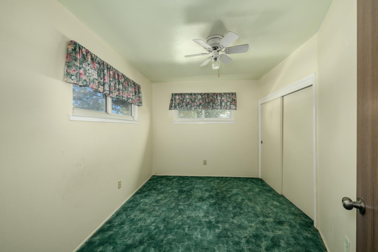 Empty bedroom with green carpet, floral window valances, and sliding closet doors.