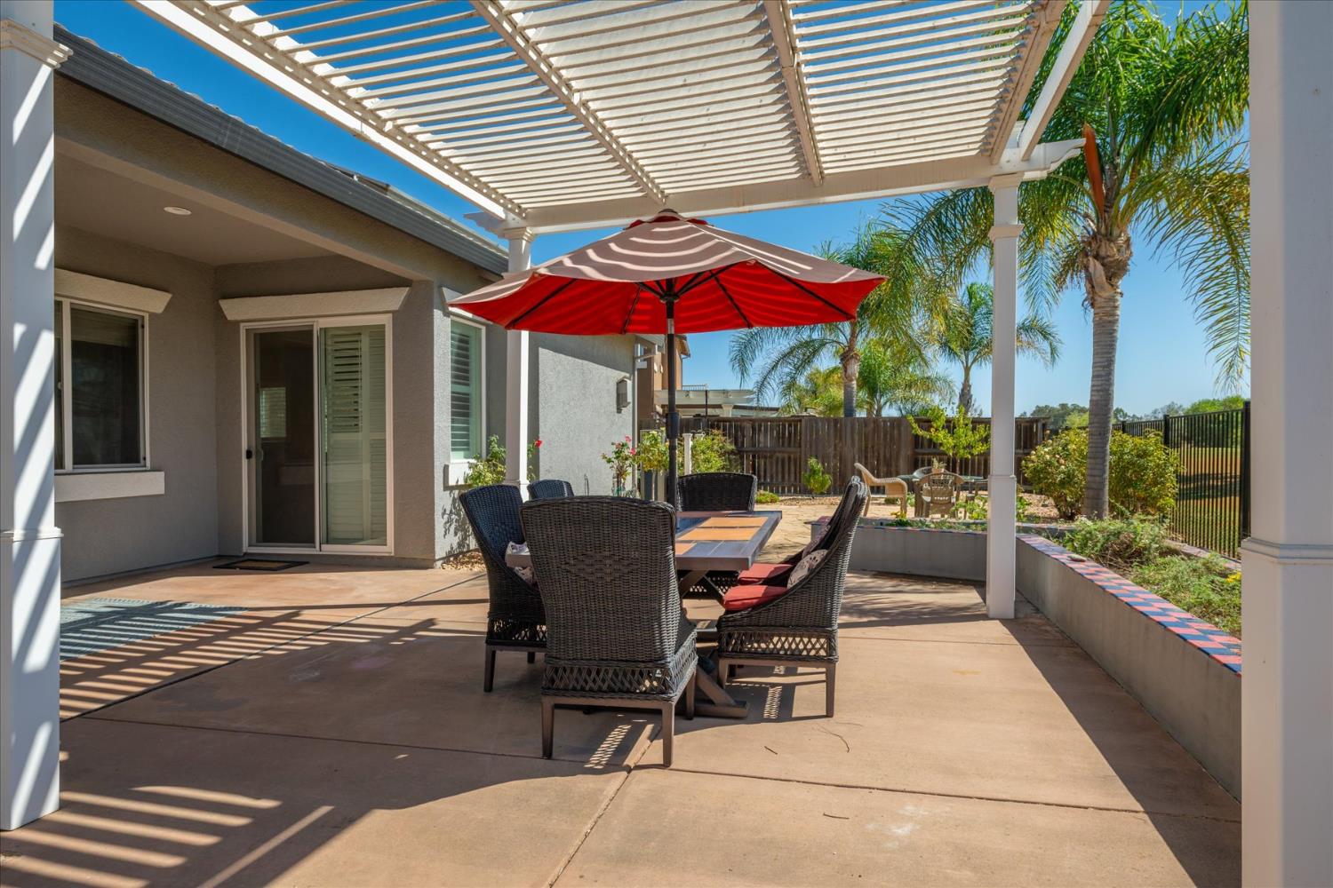 Covered patio with pergola, outdoor dining set, red umbrella, and palm trees.