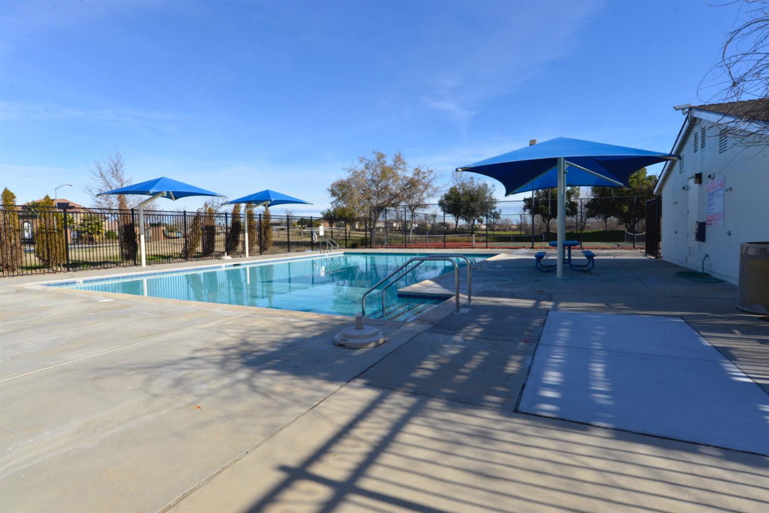 Outdoor swimming pool with a fenced deck, blue shade canopies, and picnic seating.