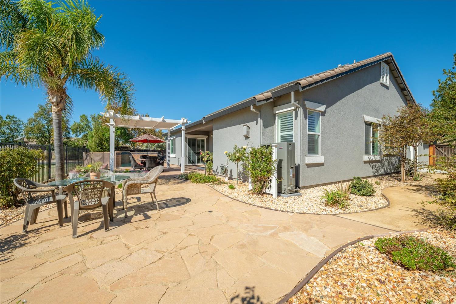 Back patio with flagstone paving, outdoor dining furniture, white pergola, and palm tree beside the gray stucco home.