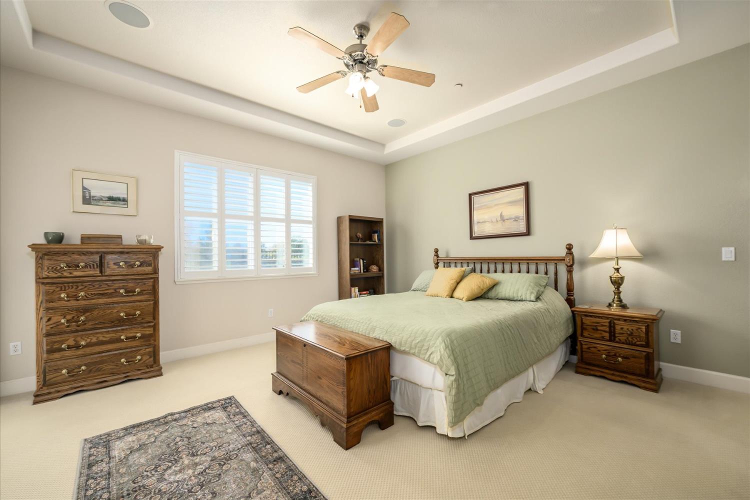Furnished bedroom with a wooden bed, plantation shutters, and a tray ceiling with ceiling fan.