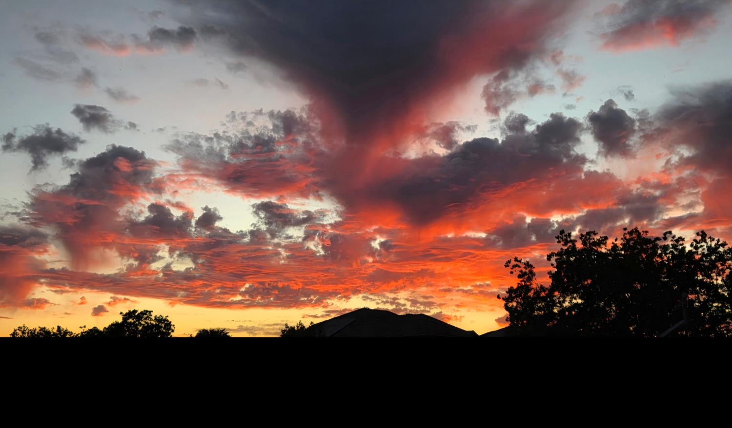 Fiery sunset clouds above silhouetted trees and rooftops.
