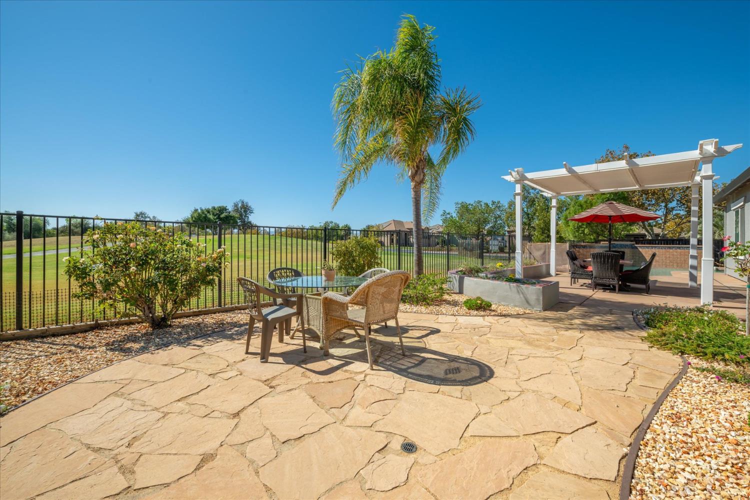 Flagstone backyard patio with a palm tree, pergola, outdoor seating, and an open green beyond the fence.