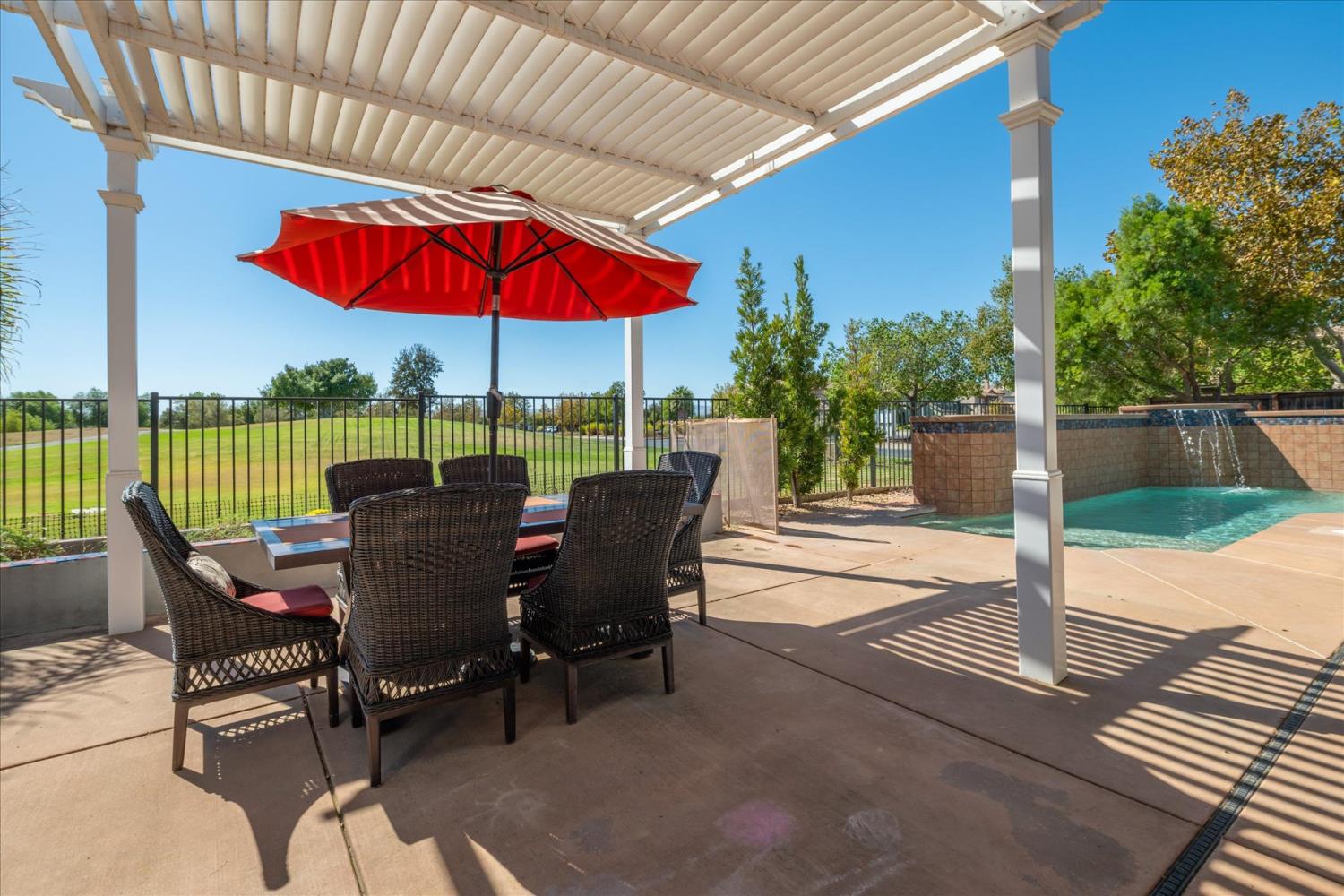 Outdoor patio with wicker seating beneath a pergola, a red umbrella, and a pool with a waterfall.