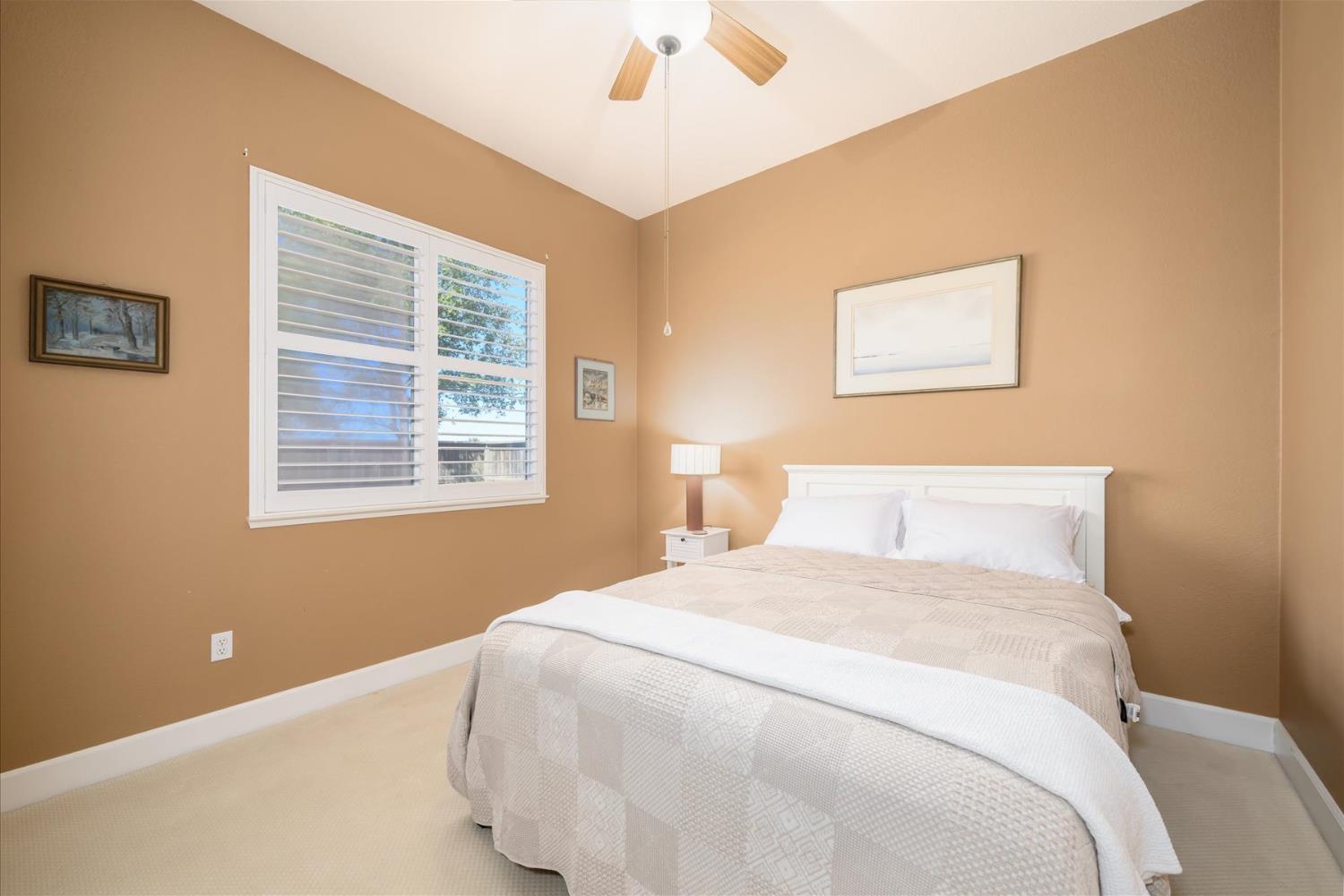 Bedroom with a white bed, tan walls, shuttered window, and ceiling fan.