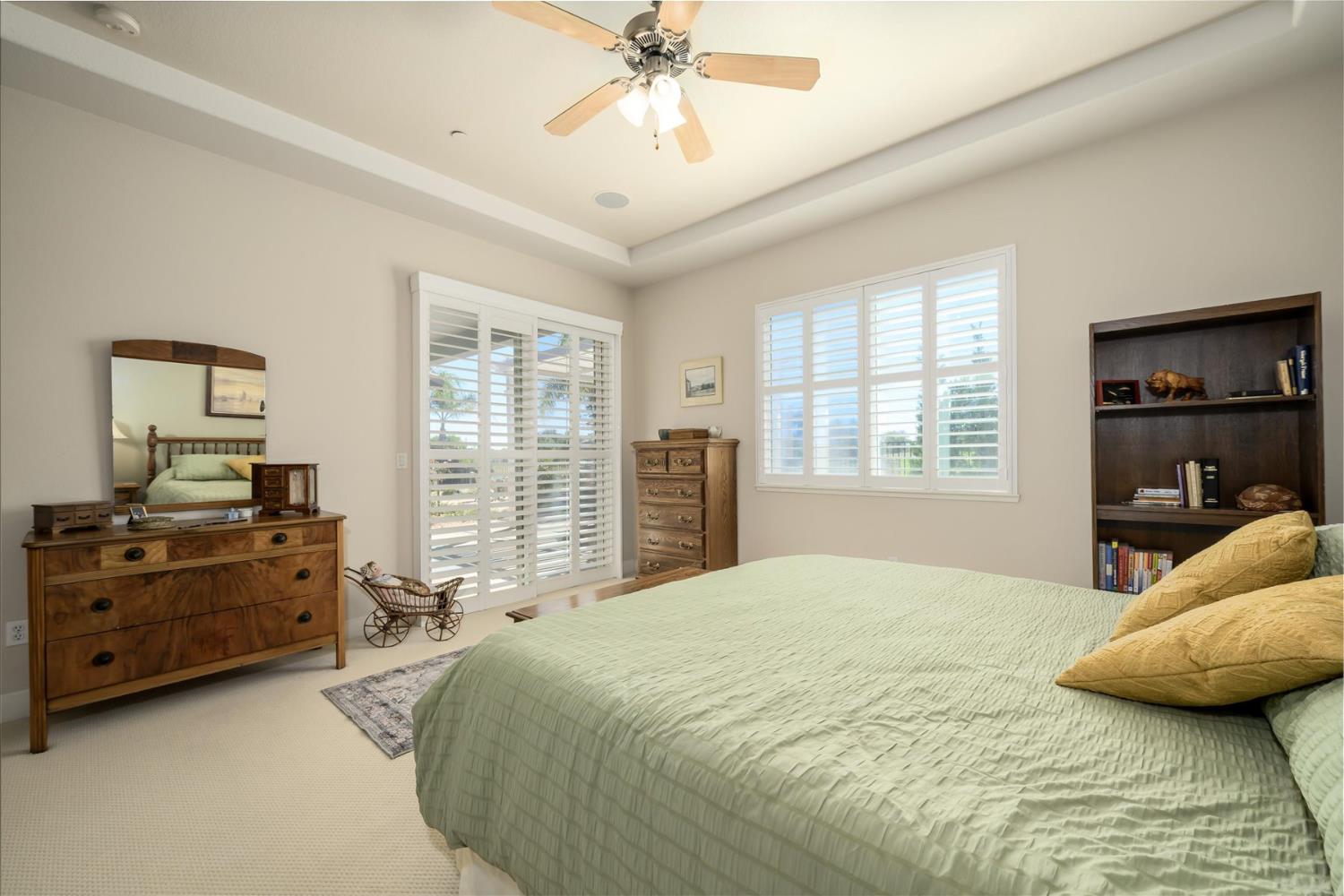 Bedroom with a green-quilted bed, plantation shutters, and a tray ceiling with a fan.