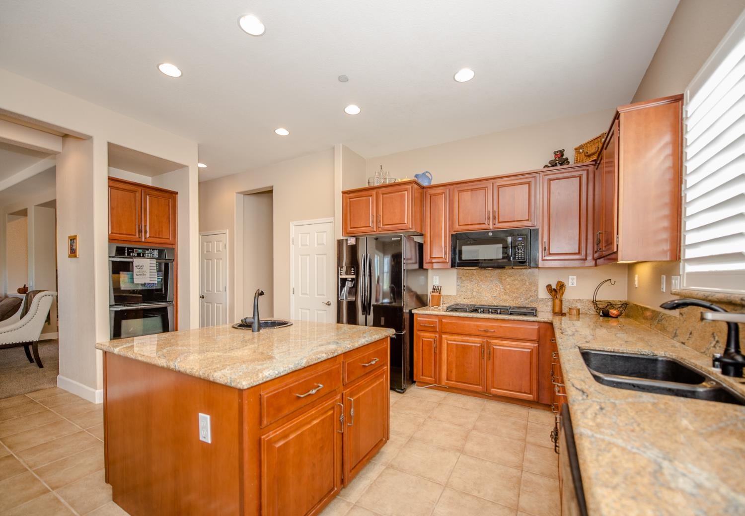 Kitchen with a large granite island, reddish wood cabinetry, stainless steel appliances, and a shuttered window.