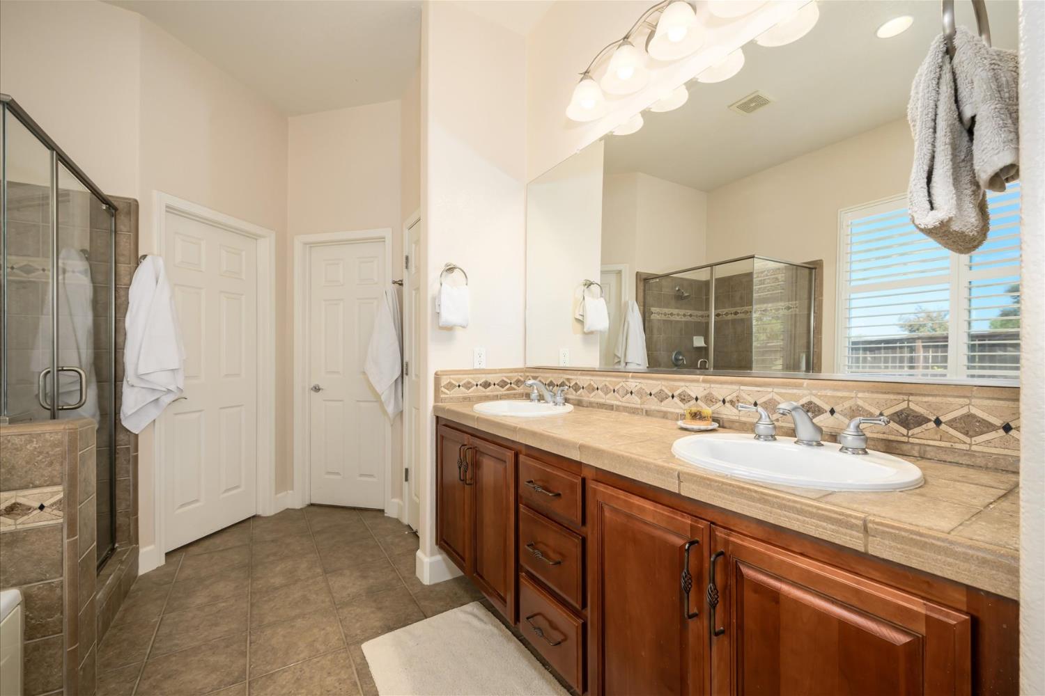 Bathroom with a double-sink wood vanity, large mirror, tiled shower, and shuttered window.