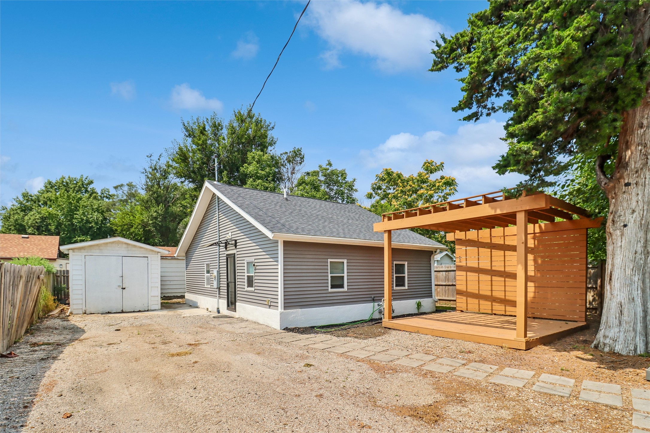 Gray-sided house with a detached garage and a large wooden pergola in the gravel backyard.