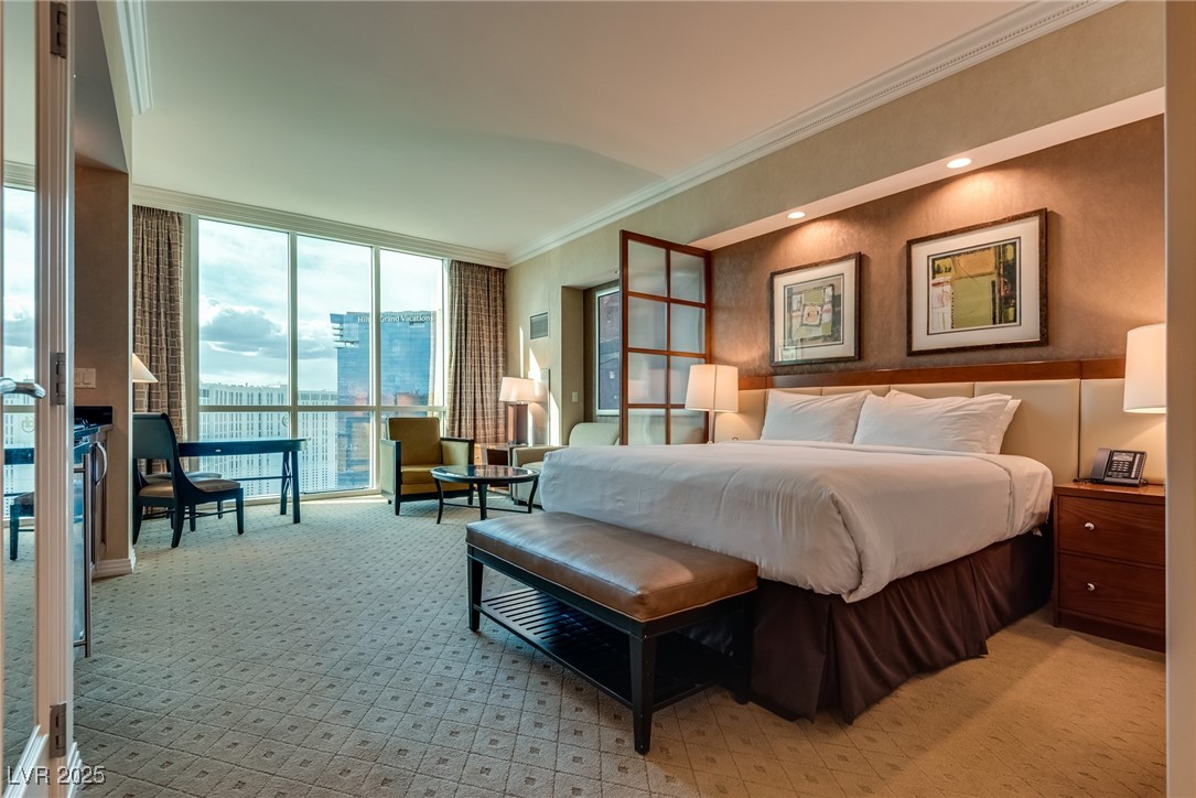 Furnished bedroom with a king bed, leather bench, and floor-to-ceiling windows overlooking high-rise buildings.