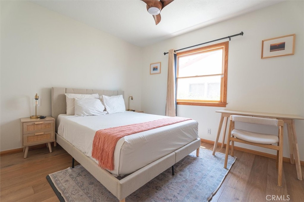 Staged bedroom with a beige upholstered bed, coral throw, and wood desk beside a bright window.
