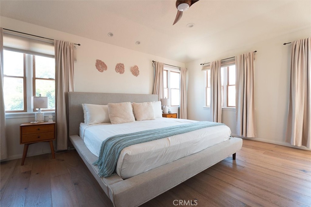 Staged bedroom with a large upholstered bed, pale blue throw, multiple curtained windows, and wood flooring.