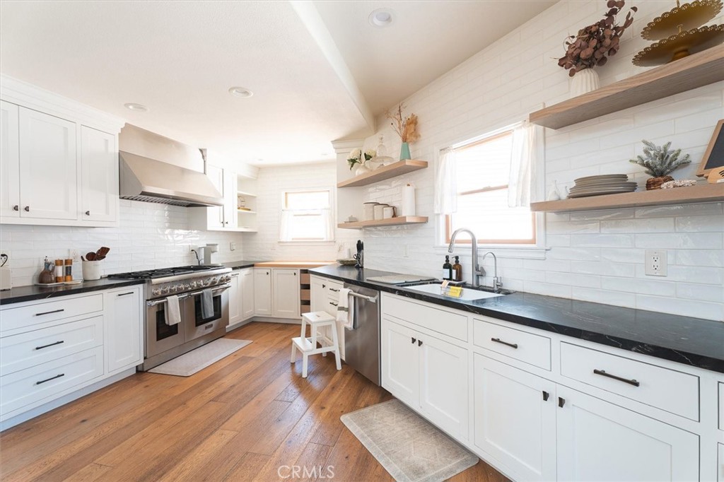 Bright white kitchen with black counters, stainless steel appliances, open shelves, and warm wood floors.