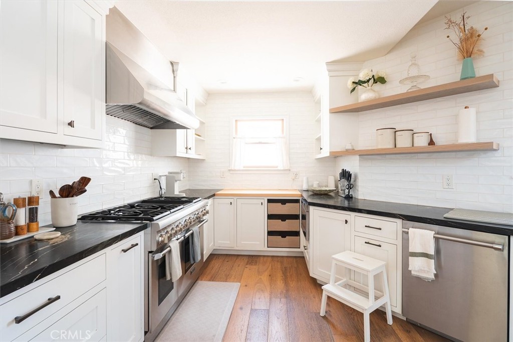 Bright white kitchen with dark countertops, stainless steel appliances, and open wood shelves.