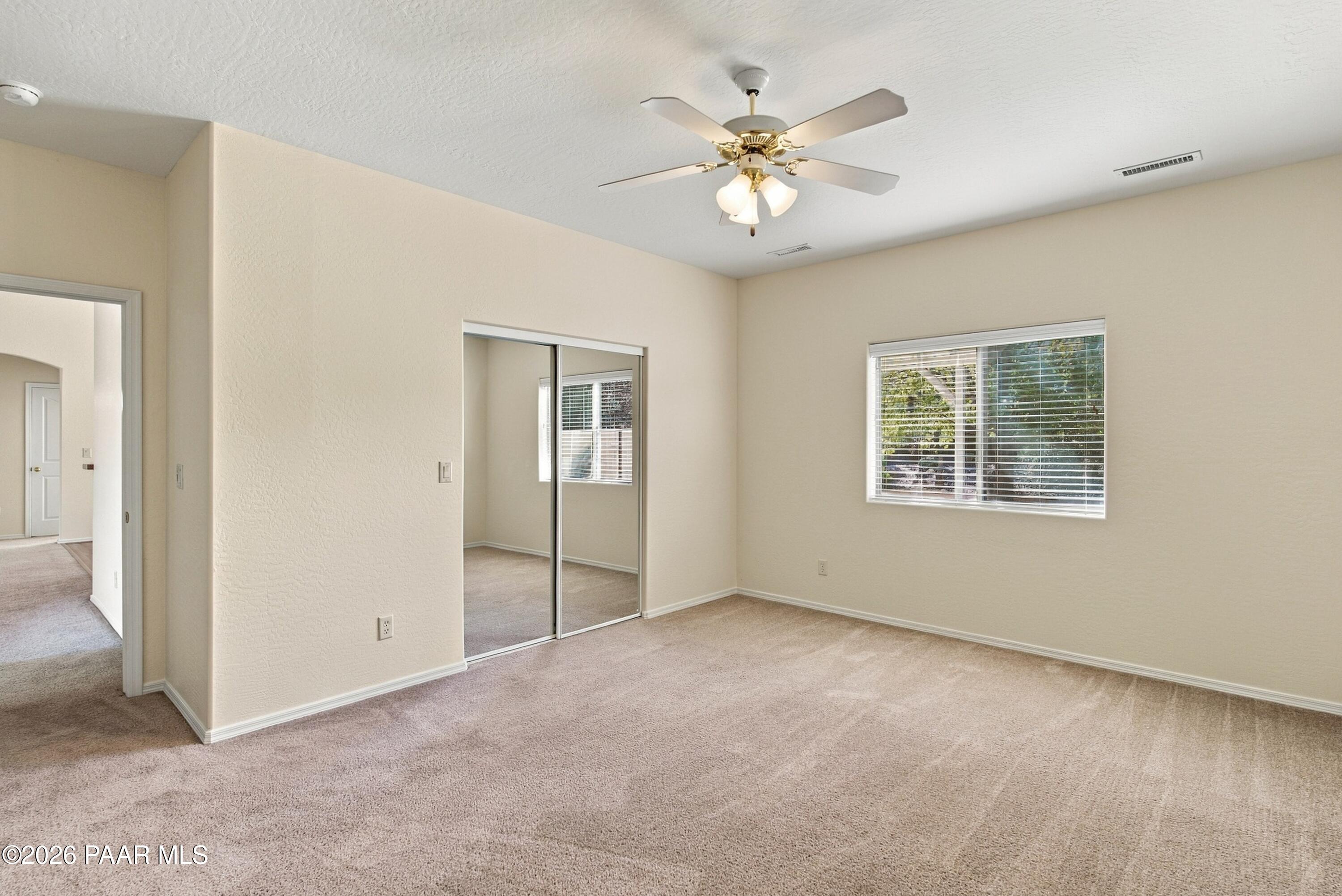 Empty bedroom with mirrored closet doors, beige carpet, a window with blinds, and a ceiling fan.