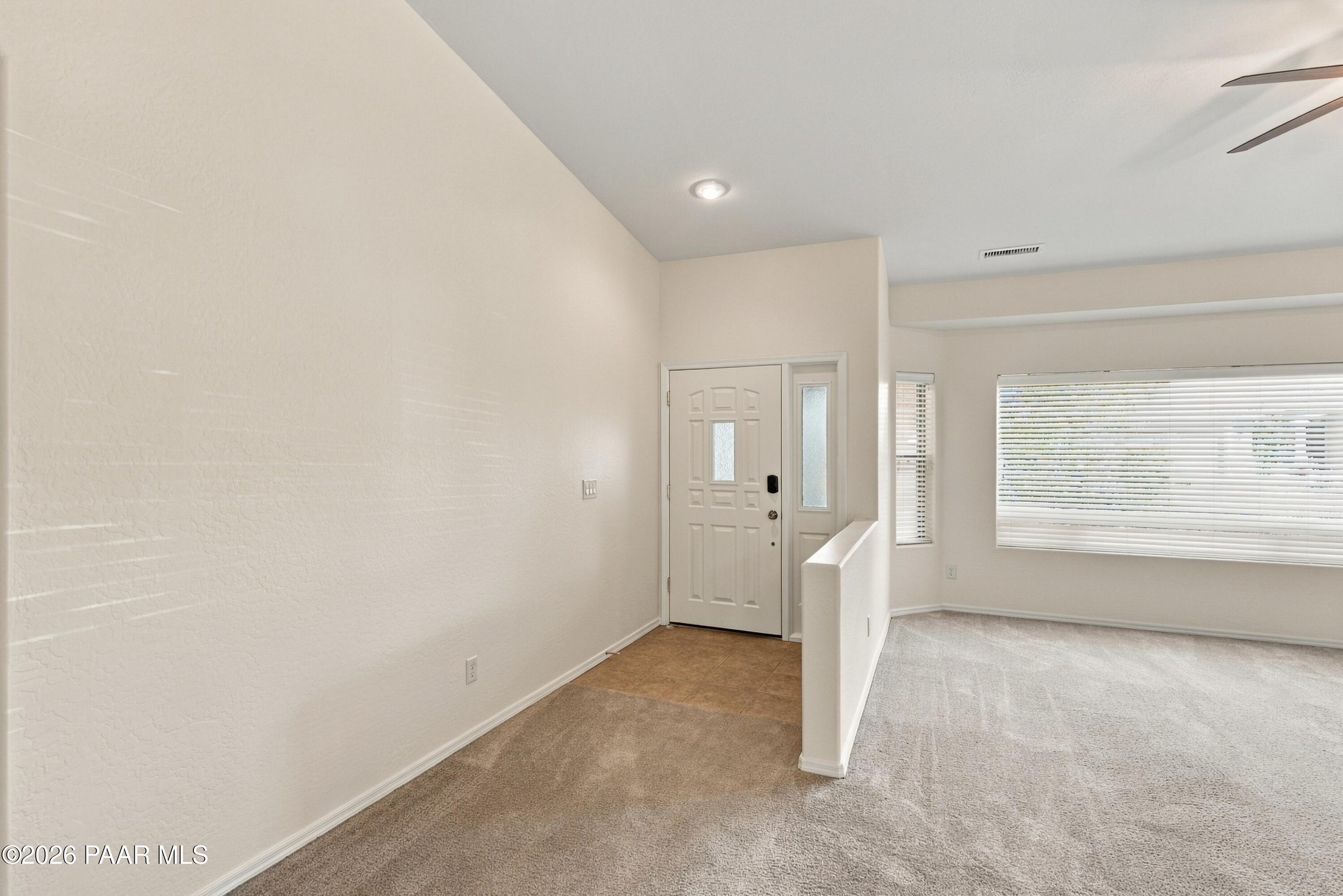Entry foyer opening to a carpeted living area, with a paneled front door and large windows with blinds.