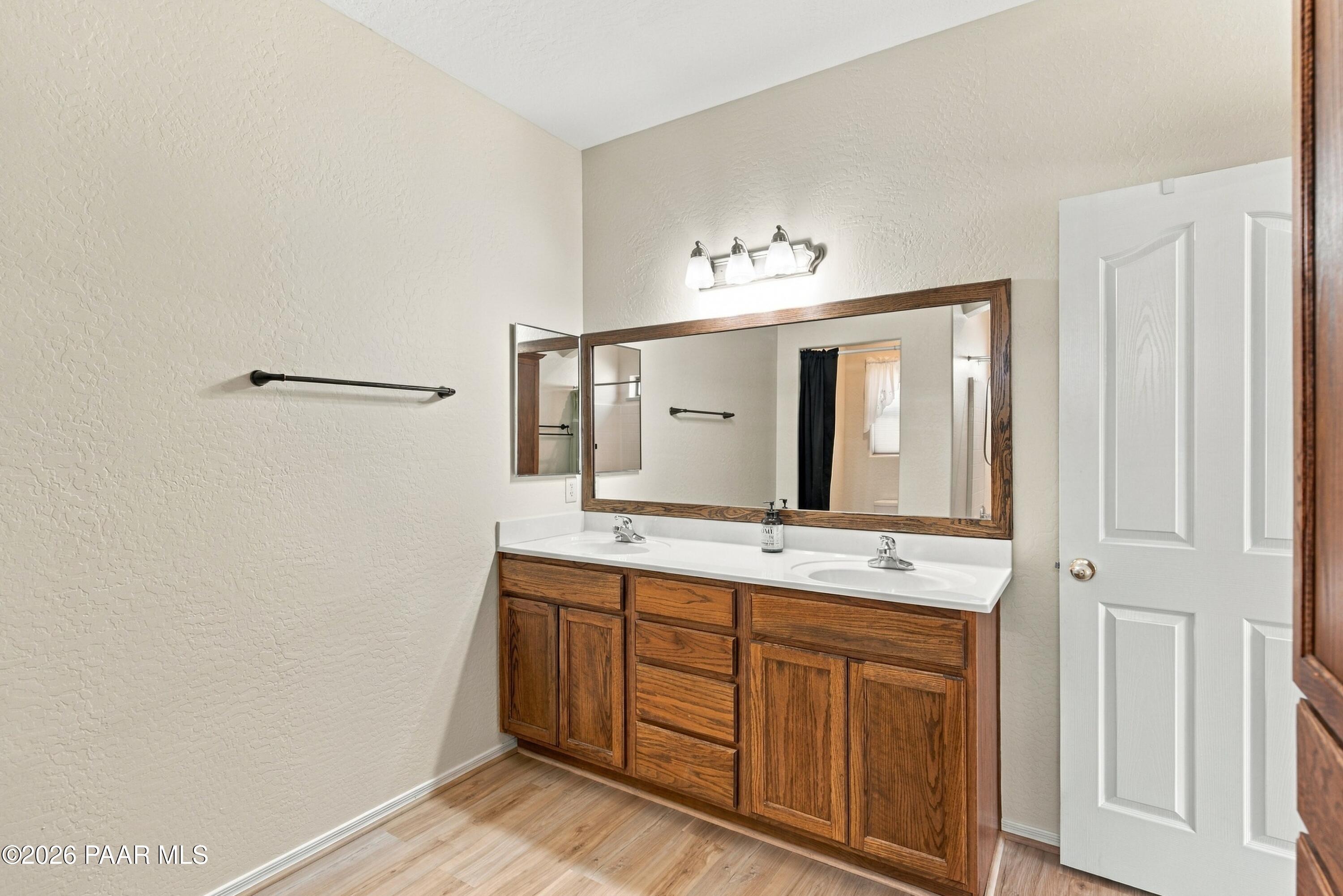 Bathroom with a double-sink wood vanity, broad framed mirror, and light wood-look flooring.