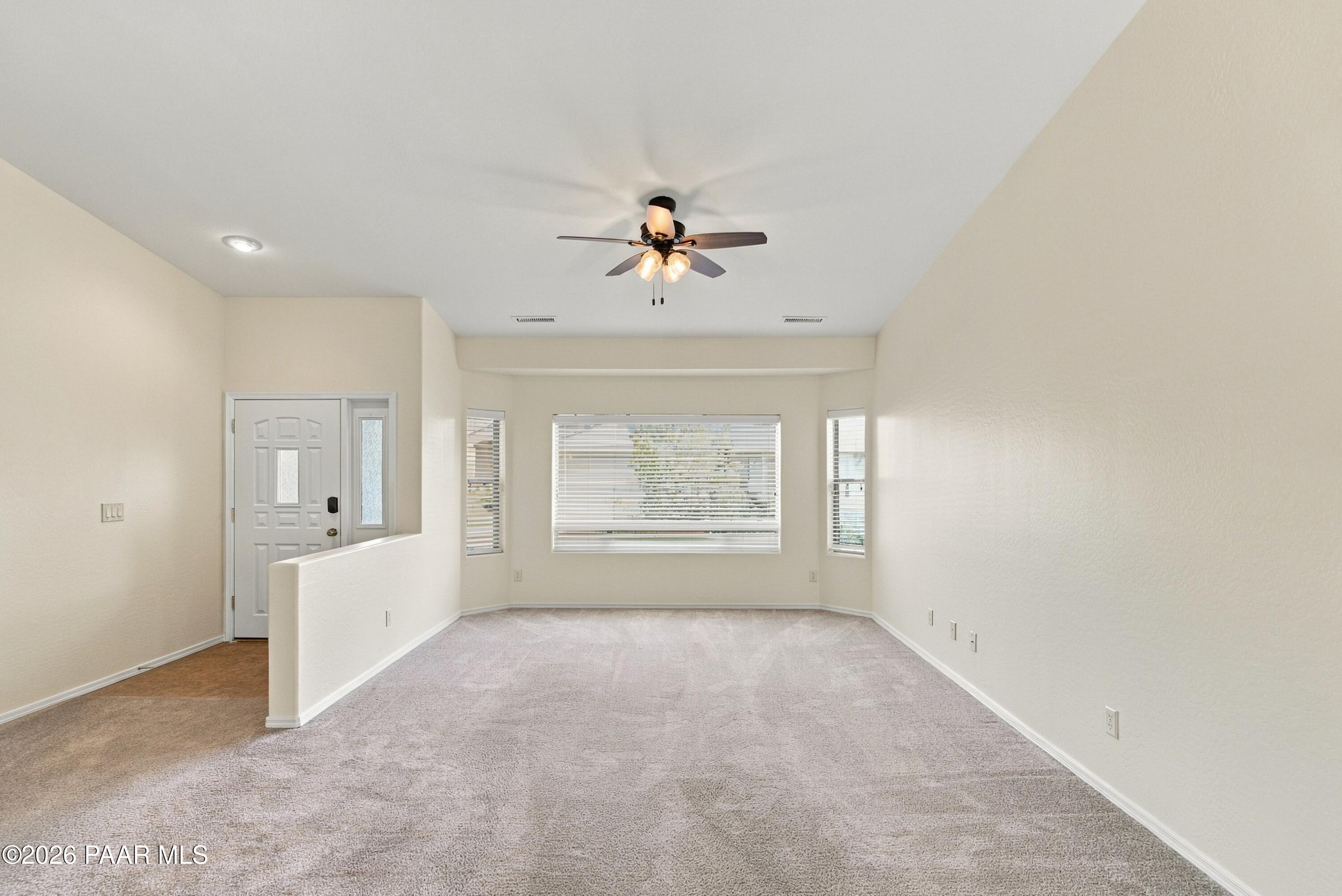 Empty living room with light carpet, ceiling fan, and wide window with blinds.